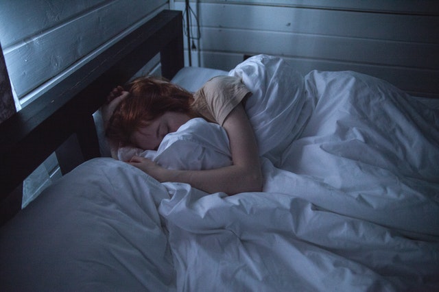 Žena s ryšavými vlasmi leží zakrytá paplónom v posteli 